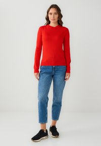 Pull rouge à manches longues en tricot lisse, assorti à un jean bleu clair et des baskets noires. Design simple sans motifs proéminents.