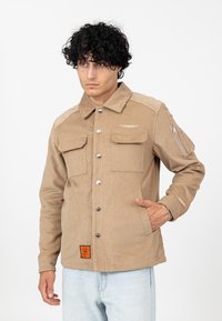 Beżowa kurtka z korduroyu z przodem na guziki, dwiema kieszeniami na piersi, kieszenią na suwak na rękawie oraz pomarańczową metką u dołu. Luźny krój.