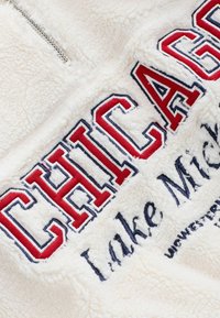 Fehér plüss pulóver, piros és kék hímzett "CHICAGO" felirattal a tetején és "Lake Michigan" írással alatta. Ezüst cipzáras zárás.
