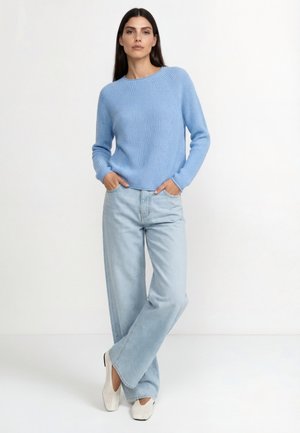 Nő világoskék kötött pulóverben, széles szárú világos farmernadrágban és fehér szőtt slip-on cipőben, zsebébe tett kézzel áll.