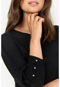 Maglietta nera a maniche lunghe con texture a costine e tre bottoni decorativi su ciascun polsino, caratterizzata da un classico scollo a girocollo.
