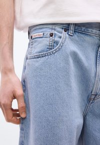 Ανοιχτό μπλε τζιν παντελόνι με μια μικρή ετικέτα Calvin Klein κοντά στην μπροστινή τσέπη, φορεμένο με ένα λευκό πουκάμισο που είναι μπροστά μέσα και με ορατό αριστερό χέρι.