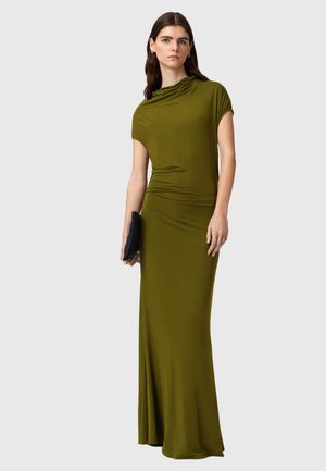 Vrouw draagt een olijfgroene, vloerlange, nauwsluitende jurk met korte mouwen, houdt een zwarte clutch vast tegen een effen lichte achtergrond.