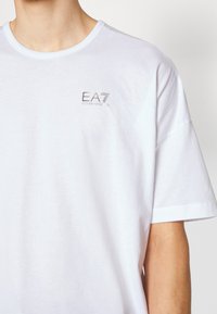 Camiseta blanca de algodón con cuello redondo, que presenta un pequeño logo "EA7" en gris bordado en el lado izquierdo del pecho. Manga corta y corte relajado.