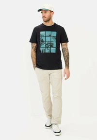 Sort t-shirt med turkis grafisk design med firkanter og tekst, parret med lys beige bukser og hvide sneakers.