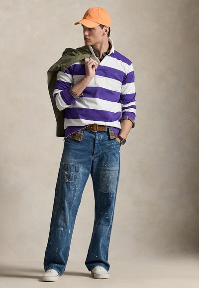 Fehér és lila csíkos hosszú ujjú ing, vállra dobott zöld dzseki, kék, bő nadrág festékfoltokkal, narancssárga sapka, fehér tornacipő.