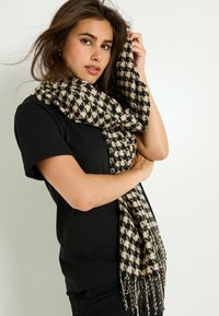 Bufanda de estampado de pata de gallo en negro y beige, hecha de un tejido suave con bordes desflecados, drapeada alrededor del cuello del modelo. Diseño texturizado y ligero.