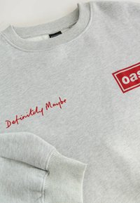 Vaaleanharmaa collegepaita, jossa on punainen teksti "Definitely Maybe" ja punainen reunuslaatikko, jossa on osittain näkyvissä sana "oas" etupuolella.
