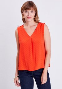 Top arancione senza maniche con scollo a V, tessuto testurizzato e dettagli con lacci regolabili sulle spalle. Abbinato a jeans scuri.