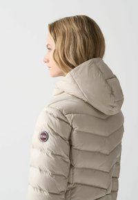 Ljusbeige pufferjacka med sydda horisontella paneler, huva och en rund logobadge på ärmen. Slätt, glänsande material.