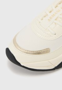 Weiße Sneaker mit goldenen Akzenten, einem Obermaterial aus strukturiertem Stoff, Schnürverschluss und einer schwarzen Gummisohle. Verfügt über minimalistische Designdetails.