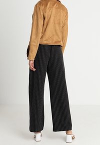 Veste en daim marron associée à un pantalon large à rayures noires et des sandales à talons bloc blanches, vue de dos.