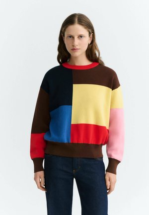 Femme portant un pull patchwork multicolore avec des sections marron, rouge, bleue, jaune et rose, associé à un jean bleu foncé, debout.