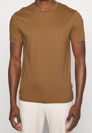 Homme portant un t-shirt marron uni à manches courtes et un pantalon beige clair, debout devant un fond neutre.