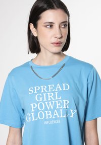 Camiseta de algodón azul claro con texto en blanco en negrita: "DIFUNDE EL PODER FEMENINO A NIVEL GLOBAL INFLUENCER." Acentuada con un collar de cadena plateada.