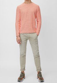 Helles korallenfarbendes Strickpullover mit langen Ärmeln und rundem Halsausschnitt, kombiniert mit hellbeigen Slim-Fit-Hosen und grünen sowie orangen Sportschuhen.