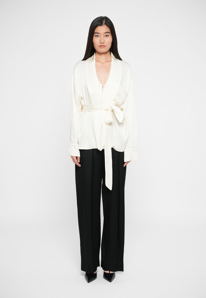 Blusa avvolgente crema con scollatura a V profonda, vita cintata e maniche lunghe, abbinata a pantaloni neri a vita alta con gamba ampia e scarpe nere a punta.