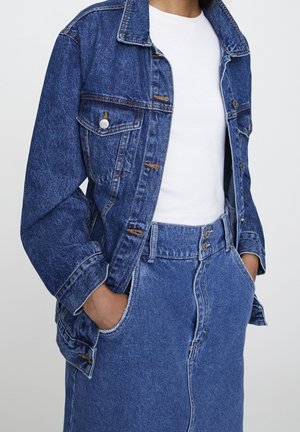 Personne portant une veste en jean bleue et une jupe en jean taille haute assortie, les mains dans les poches, sur un haut blanc côtelé.