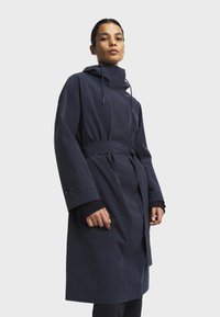 Laivastonsininen vedenkestävä trench-coat, jossa on istuva siluetti, vyötärölle sidottu vyö, korkea kaulus ja sivutaskut. Ominaisuuksiin kuuluvat leveät hihat ja minimalistinen muotoilu.