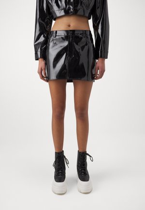 Model iført sort skinnende vinyl cropped jakke og matchende mini nederdel med sort-hvide platform snørestøvler, stående mod hvid baggrund.