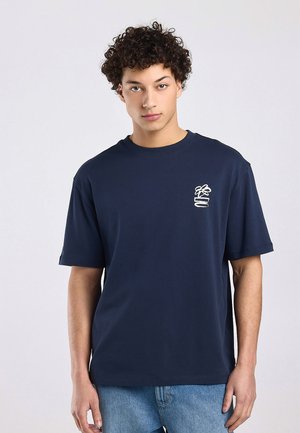 Joven con cabello rizado que lleva una camiseta azul marino con un pequeño gráfico blanco en el lado izquierdo del pecho y jeans azul claro, de pie mirando al frente.