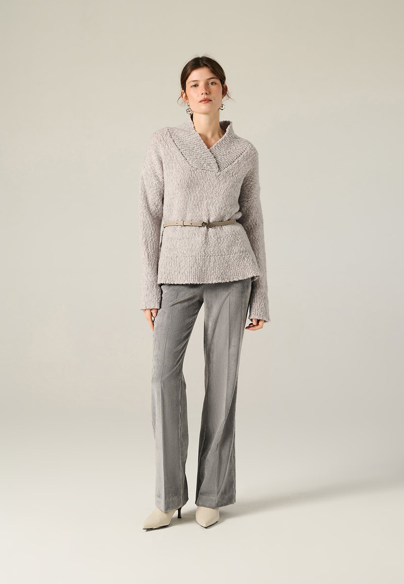 Maglione grigio chiaro con texture e colletto ampio, abbinato a pantaloni grigi a righe e décolleté beige a punta, accentuato in vita da una cintura.