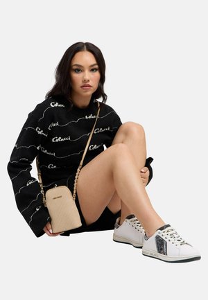 Kvinde siddende iført sort langærmet top, sorte shorts, hvide sneakers og beige crossbody taske mod en ensfarvet hvid baggrund.