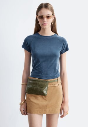 Jeune femme aux longs cheveux portant un haut bleu à manches courtes, une mini-jupe beige, des lunettes de soleil, des bijoux en or et un sac banane vert olive.