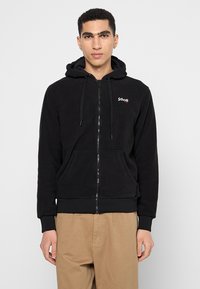 Schwarzer Kapuzenpullover mit Reißverschluss aus weichem Fleece-Material, mit einer Fronttasche, Kordelzügen und einem kleinen roten Logo auf der linken Brust.