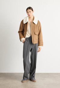 Bruine cropped jacket met witte faux fur kraag, gecombineerd met grijze wijde broek en grijze schoenen. Zachte texturen en functionele ontwerpelementen.