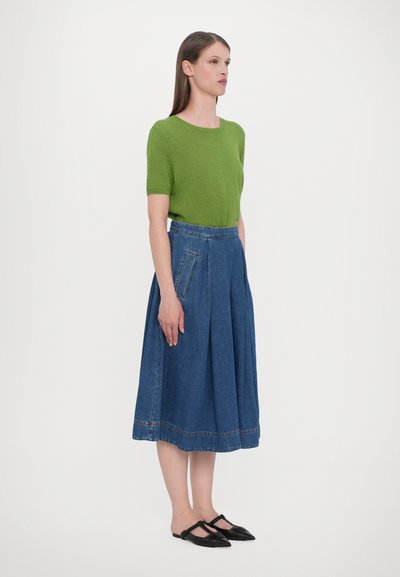 Groene gebreide top met korte mouwen, hoograitzijde blauwe denim geplooide rok, middenkuitlengte, zwarte platte schoenen met textuurdetails.