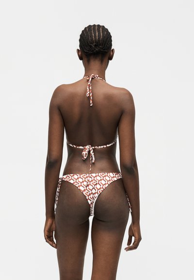 Γυναίκα με πλεκτά μαλλιά που φορά μπικίνι με καφέ και λευκό μοτίβο, όρθια με την πλάτη προς το μέρος μας σε απλό φωτεινό φόντο.