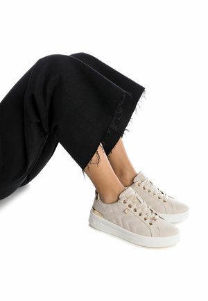 Zapatillas beige con parte superior texturizada, acentos de hardware dorado y suela blanca. Pantalones de pierna ancha negros con dobladillo deshilachado visible en los tobillos.