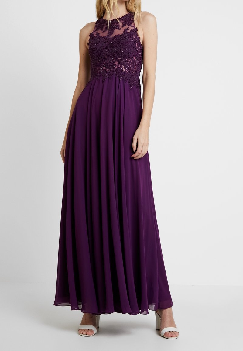 Fioletowa wieczorowa suknia z przezroczystym, koralikowym gorsetem i zwiewną spódnicą z szyfonu. Posiada wysoki dekolt i sięga do kostek, zestawiona z sandałkami na cienkich paskach.