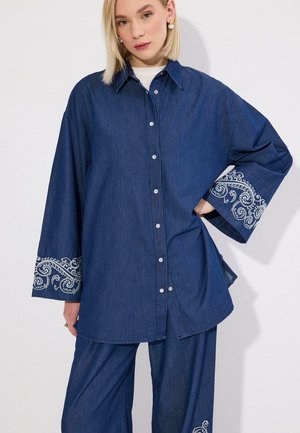 Blond kvinde iført løs mørkeblå denimskjorte med hvidt broderi på brede ærmer og matchende bukser med vide ben mod hvid baggrund.