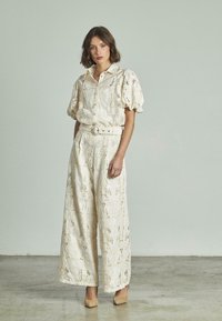 Completo bianco sporco composto da una camicia con maniche voluminose e chiusura a bottoni e pantaloni a gamba larga con intricati motivi ricamati.