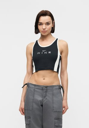 Tânără cu păr scurt și închis la culoare, purtând un top scurt Nike alb-negru și pantaloni cargo gri, stând în fața unui fundal deschis, simplu.