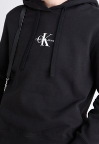 Felpa con cappuccio nera con logo bianco di Calvin Klein Jeans. Realizzata in tessuto morbido con lacci e polsini a costine.