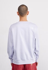 Osoba s krátkými vlasy má na sobě světle fialovou mikinu The North Face a červené kalhoty, stojí zády k objektivu před jednoduchým bílým pozadím.