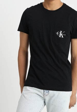 Hombre que lleva una camiseta negra de Calvin Klein Jeans con un logo en el bolsillo del pecho combinada con vaqueros azul claro sobre un fondo liso.