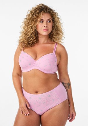 Krøllet kvinde iført pink broderet bh med blomster og matchende højtaljede trusser, drejet let mod venstre på hvid baggrund.