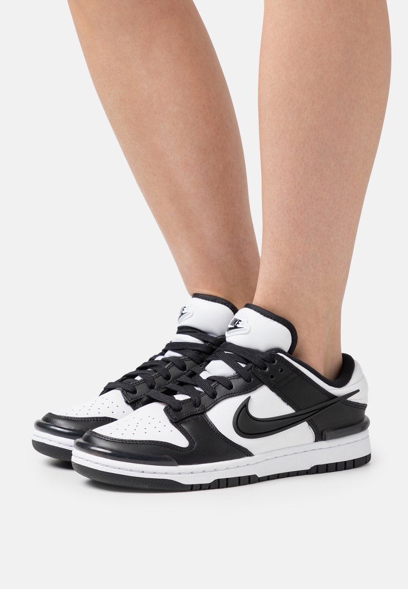 Pieds portant des baskets Nike noires et blanches avec des lacets noirs, présentés sur un fond clair uni.