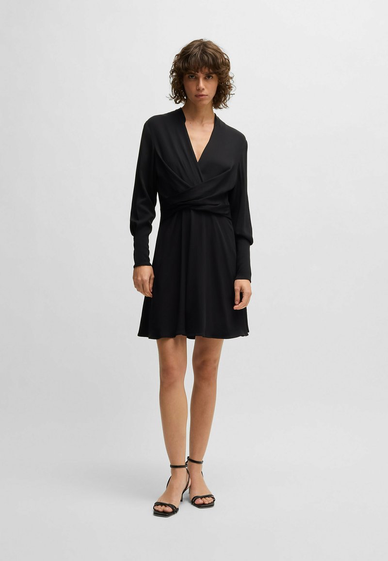 Black wrap dress with long sleeves, V-neckline, and fitted waist. Soft, smooth fabric with a flared hem, paired with black strappy sandals.