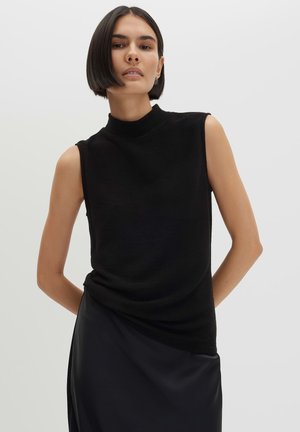 Femme aux cheveux courts et foncés portant un haut noir sans manches et une jupe noire sur un fond clair uni.