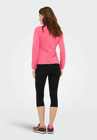 Maglia rosa a maniche lunghe, leggings neri corti e sneaker marroni con dettagli rosa. Tessuto liscio, vestibilità atletica e colori a contrasto.