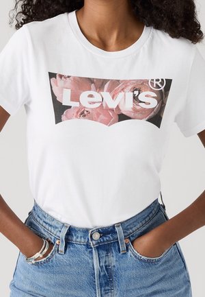 Vrouw draagt een wit Levi's T-shirt met roze bloemenlogo, gecombineerd met hoog getailleerde blauwe spijkerbroek en armbanden aan de linkerpols.
