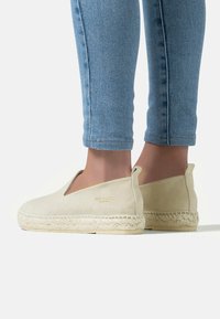 Beige espadrille in soft suede with a braided jute sole and cutout sides, featuring a minimal logo detail. Worn with blue skinny jeans.
