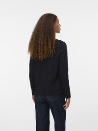 Γυναίκα με μακριά σγουρά μαλλιά, φορώντας μαύρη μπλούζα με μακριά μανίκια και σκούρα μπλε τζιν, όρθια, στραμμένη μακριά σε ένα απλό λευκό φόντο.