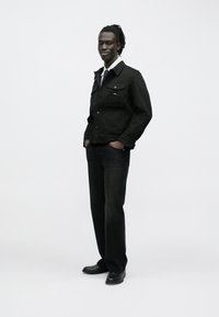 Čierna rifľová bunda s golierom a detailmi na gombíky, v kombinácii s tmavými rifľovými nohavicami a čiernymi topánkami. Hladká textúra, priliehavý strih.