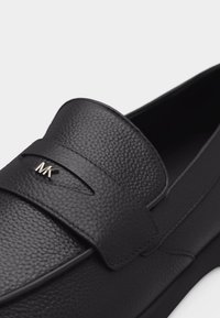 Μαύρο δερμάτινο μοκασίνι με υφή και λουράκι που φέρει μικρό μεταλλικό λογότυπο "MK" στο μπροστινό πάνω μέρος του παπουτσιού.